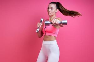 jovem confiante em roupas esportivas exercitando com halteres contra fundo rosa foto