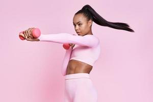 confiante jovem africana se exercitando com halteres em pé contra um fundo rosa foto