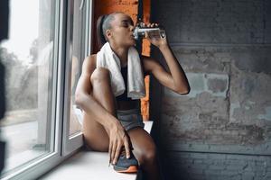 Atraente jovem africana bebendo água enquanto está sentado no peitoril da janela no ginásio foto