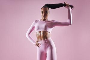 mulher africana atraente em roupas esportivas, ajustando o cabelo contra o fundo rosa foto