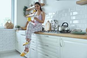 jovem relaxada usando telefone inteligente e tomando café enquanto está sentado no balcão da cozinha foto