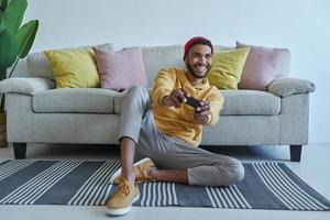 animado jovem africano jogando videogame enquanto está sentado no chão em casa foto