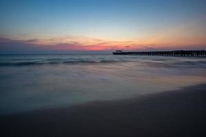 a sombra da ponte do porto. o pôr do sol, o céu laranja, o mar e a praia refletem a luz laranja. praia de natai na província de phang nga, tailândia foto