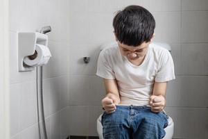 menino asiático sentado no vaso sanitário segurando papel de seda - conceito de problema de saúde foto