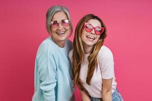 feliz mãe e filha adulta usando óculos funky em pé juntos contra o fundo rosa foto