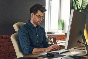 jovem bonito usando computador enquanto está sentado em seu local de trabalho no escritório foto