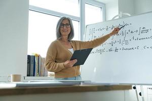 alegre mulher sênior ensinando matemática enquanto aponta o quadro branco na sala de aula foto