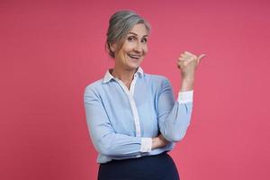 mulher sênior feliz apontando para longe em pé contra um fundo rosa foto