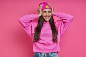 mulher jovem surpresa na camisa com capuz, segurando a cabeça nas mãos contra o fundo rosa foto