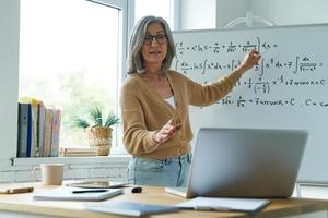 mulher madura ensinando matemática enquanto aponta o quadro branco e olhando para o laptop foto