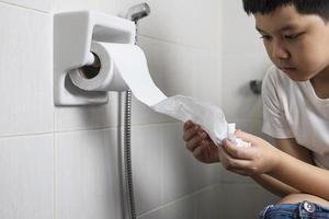 menino asiático sentado no vaso sanitário segurando papel de seda - conceito de problema de saúde foto