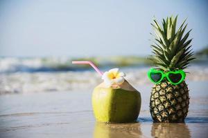 coco fresco e abacaxi colocam óculos adoráveis de sol na praia de areia limpa com fundo de ondas do mar - frutas frescas com conceito de fundo de férias de sol de areia do mar foto