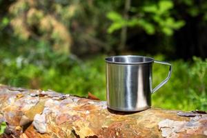 caneca turística de metal com bebida quente fica no tronco de árvore na floresta. foto