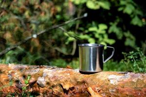 caneca turística de metal com bebida quente fica no tronco de árvore na floresta. foto