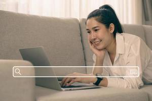 alegre sorridente mulher asiática usando laptop com ícone de pesquisa enquanto está sentado no sofá na sala de estar. ela usa seu laptop para pesquisar e fazer compras online. foto