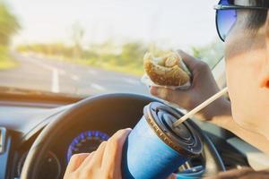 homem está comendo perigosamente cachorro-quente e bebida gelada enquanto dirige um carro foto