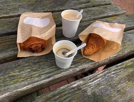 copos de papel com café saboroso e croissant francês no pacote de papel, lanche da manhã para viagem na velha mesa de madeira do lado de fora do parque, vista superior foto