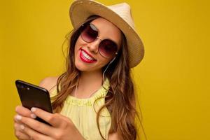 retrato de uma jovem muito sorridente com maquiagem brilhante, vestindo roupas de verão em pé isolado sobre fundo amarelo, posando com chapéu, usando smartphone foto