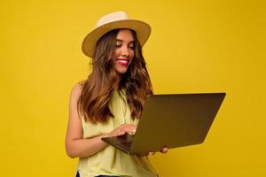 adorável jovem europeia atraente com cabelos ondulados usando chapéu e vestido brilhante de verão trabalhando com laptop isolado sobre fundo amarelo foto