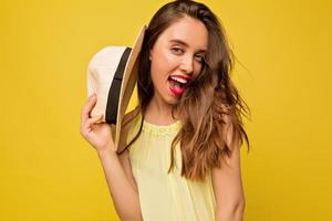 incrível mulher de cabelos compridos vestido de verão brilhante posando com emoções expressivas, ela abriu o moush e segura o chapéu perto do rosto em fundo amarelo foto