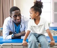 paciente infantil sentado na cama do hospital com médico africano para atendimento médico. sorridente menina feliz e saudável após exame profissional na clínica. médico, pediatra verificando, examinando criança foto