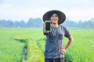 jovem agricultor inteligente milenar mostrando smartphone de tela em branco no meio do campo de arroz. tecnologia moderna para a agricultura. foto