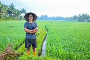 Atraente alegre jovem agricultor asiático em pé com confiança e olhando para a câmera no campo de arroz. conceito de agricultura moderna. foto