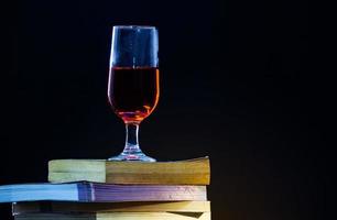 livros antigos se sobrepõem em um fundo preto e com pouca luz um copo de vinho tinto em cima. foto