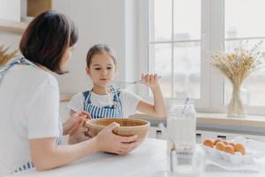 adorável criança de avental mostra bata com creme branco, usa avental, cozinha junto com a mamãe, posar na cozinha espaçosa juntos, fazer bolo para evento especial. crianças, ajuda com a casa foto