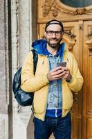 jovem viajante em roupas da moda em pé no museu tendo um olhar feliz por estar lá. macho barbudo alegre na jaqueta amarela segurando a mochila nas costas segurando o celular visitando o museu regional foto