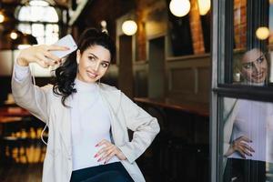 mulher morena alegre em roupas elegantes brancas segurando o telefone móvel genérico fazendo sefie contra o fundo do café aconchegante. elegante jovem feminina fazendo foto de si mesma se divertindo. tecnologia moderna