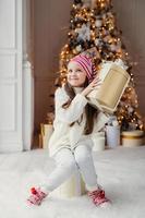 criança pequena curiosa não sabe o que está na caixa de presente, recebe presente do papai noel, usa roupas de malha quentes, senta-se contra o fundo decorado da árvore de ano novo. infância, surpresa, férias foto