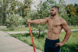 homem atlético musculoso tem treino com poses de banda de resistência elástica com corpo musculoso sem camisa, fica ao ar livre, concentrado na distância. desportista atleta tem treino no parque. estilo de vida saudável foto