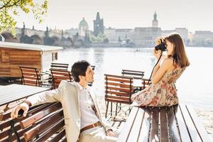 profissional linda fotógrafa feminina faz foto do marido que posa para a câmera no banco de madeira no café ao ar livre perto da bela vista panorâmica da cidade antiga. pessoas e conceito de hobby