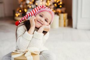 linda criança pequena posa na sala de estar, inclina-se no presente presente, tem expressão feliz, feliz em receber surpresa dos pais, passa férias no círculo familiar. feliz Natal e Feliz Ano Novo foto