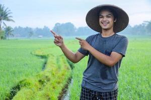 Atraente alegre jovem agricultor asiático em pé, sorrindo para a câmera e apontando o dedo para o campo de arroz. conceito de agricultura moderna. foto