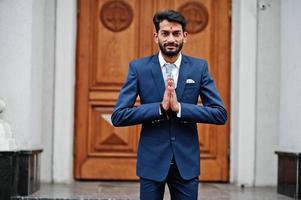 homem indiano de barba elegante com bindi na testa, use terno azul posou ao ar livre contra a porta do prédio e mostre o sinal de mãos namastê. foto