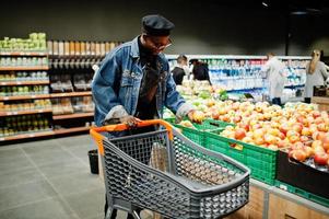 homem afro-americano casual elegante na jaqueta jeans e boina preta na seção orgânica do supermercado com carrinho de compras. foto