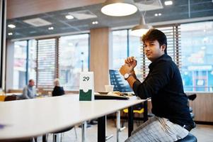 homem indiano elegante sentado no café fast food contra seu laptop e comendo batatas fritas. foto