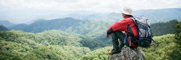 jovem caminhadas masculinas no topo da rocha, mochila homem olhando para o belo vale da montanha à luz do sol no verão, paisagem com esporte homem, altas colinas, floresta, céu. viagem e Turismo. foto