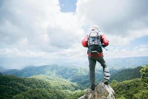 jovem caminhadas masculinas no topo da rocha, mochila homem olhando para o belo vale da montanha à luz do sol no verão, paisagem com esporte homem, altas colinas, floresta, céu. viagem e Turismo. foto