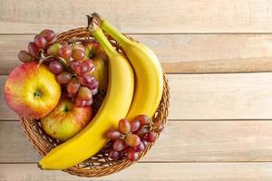 frutas maduras na cesta de vime com fundo de madeira. bananas, maçãs e uvas. conceito de colheita. foto