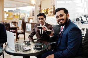 dois homens de negócios indianos de terno sentado no escritório no café, olhando para o laptop e tomando café. foto