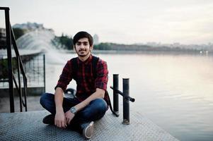 homem jovem estudante indiano na camisa quadriculada e jeans com mochila posou na cidade à noite contra fontes. foto