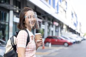 jovem estudante mulher asiática segurando café e tablet usando protetor facial para prevenir vírus foto