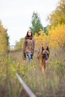 mulher jovem e atraente andando com seu cão pastor alemão na floresta de outono, perto da ferrovia - a garota está em foco foto