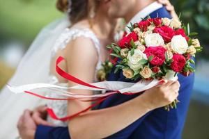 a noiva pôs as mãos nos ombros do noivo. noiva com um buquê de rosas vermelhas e brancas abraça e beija o noivo na natureza. foto