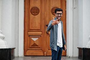 homem indiano elegante de óculos usa casual posou ao ar livre contra a porta do prédio, mostra sinal de dedos de pedra. foto