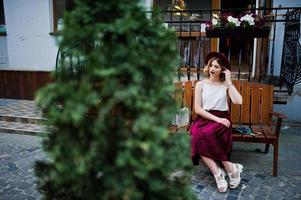 menina modelo loira elegante e bonita na saia de veludo vermelho elegante, blusa branca e chapéu, posou no banco na rua da cidade. foto