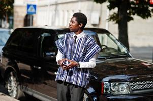 empresário africano em roupas tradicionais e óculos com celular contra carro preto suv. ricos africanos. foto
